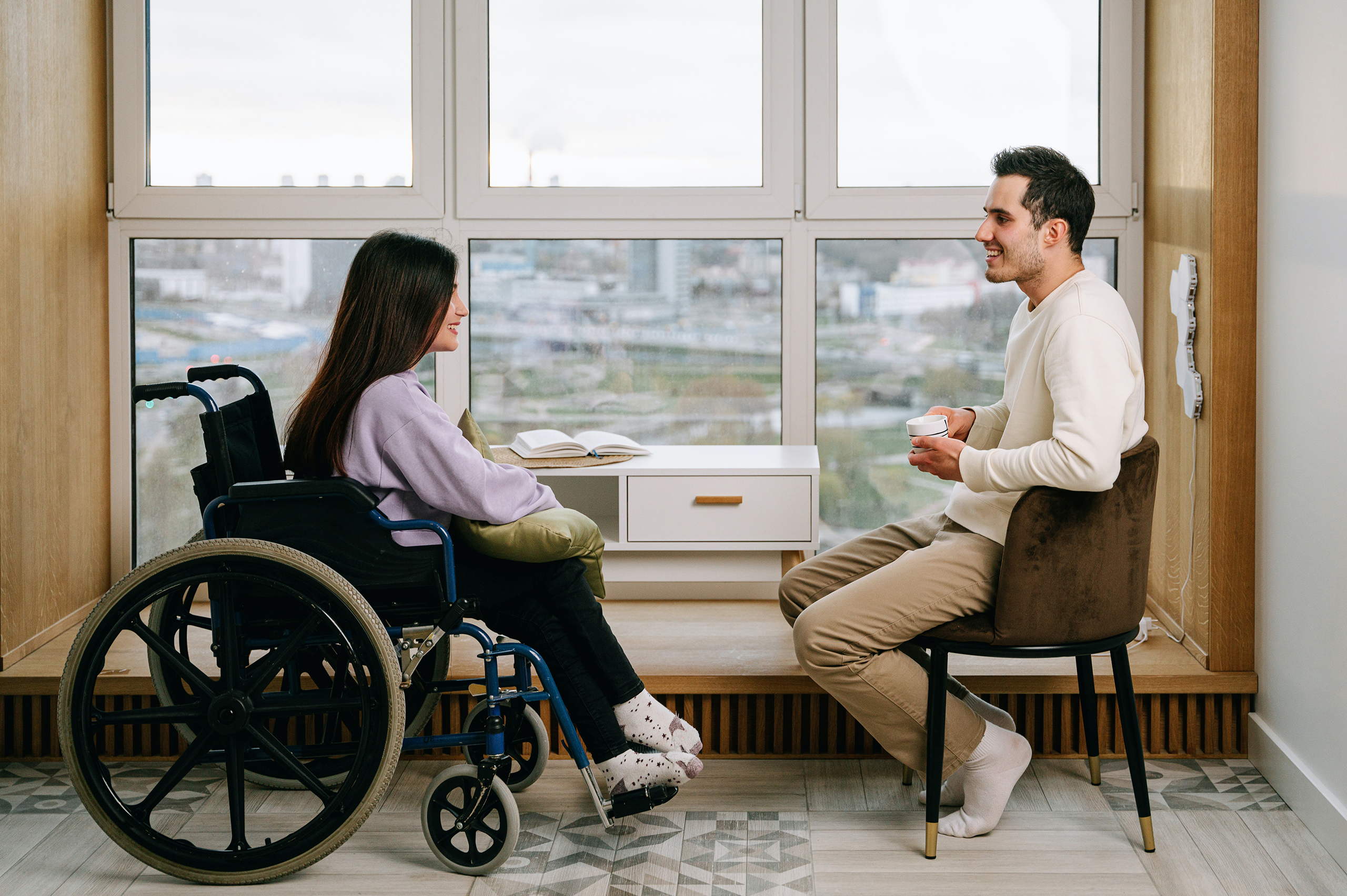 A woman in a wheelchair and a man in a chair have a conversation next to a window