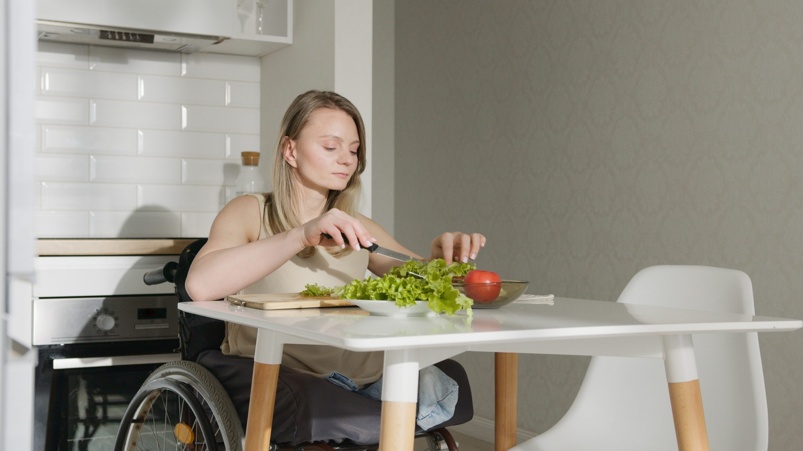 A woman in a wheelchair cuts vegetables at her kitchen table