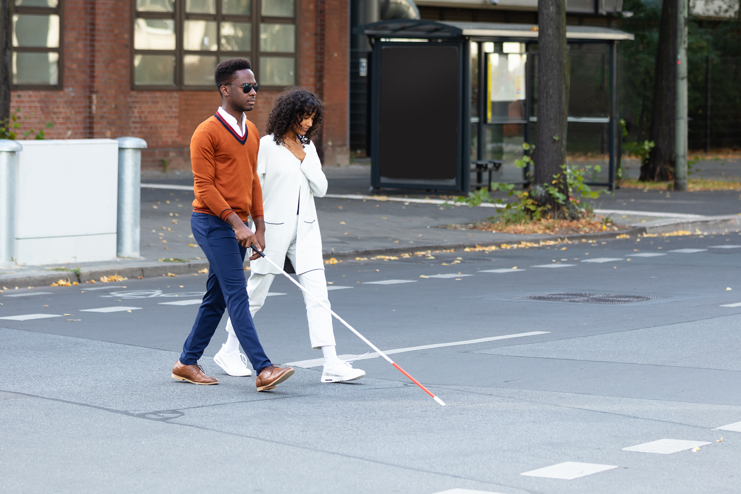 A Blind man in an orange sweater uses his cane to cross the street under the supervision of the Orientation & Mobility Specialist walking with him