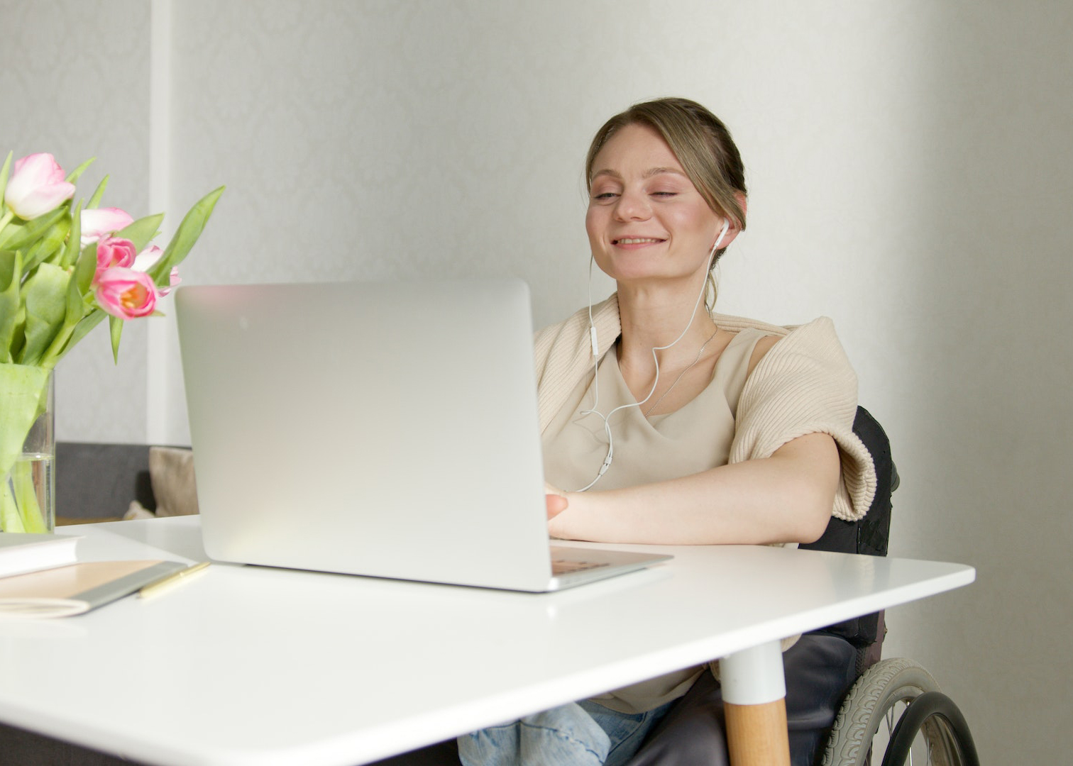 A woman in a wheelchair wearing earbuds smiles as she watches an online course on her computer